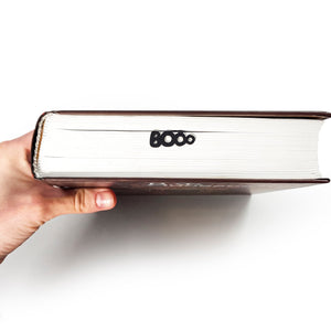 Hand holding a thick book with a black metal “BOO” bookmark visible between the pages, peeking out from the spine. Playful Halloween-themed detail.
