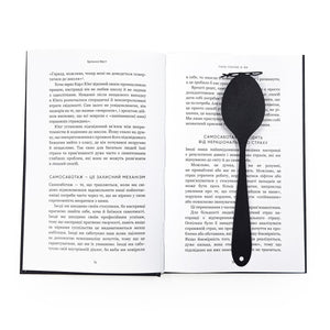 Black metal spoon-shaped bookmark placed vertically in the center fold of an open book with Ukrainian text on both pages.