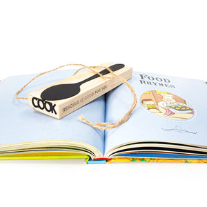 Black metal spoon-shaped bookmark displayed on a wooden block engraved with “READING IS GOOD FOR YOU” and “COOK,” placed over an open children’s book titled Food Rhymes.