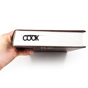Hand holding a closed book with a black metal bookmark inside; the word “COOK” is visible protruding from the book’s spine edge.