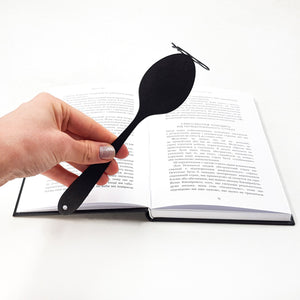 Hand holding a black metal spoon-shaped bookmark above an open book with Ukrainian text, displayed on a white background.