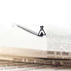 Close-up of a black teepee-shaped metal bookmark lying on an open book page, with stacked vintage-style papers in the foreground.
