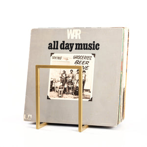 Minimalist metal vinyl record stand in gold finish holding a large collection of LPs, displayed on white background.