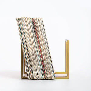 Side view of a minimalist metal vinyl record stand in gold finish holding a large collection of LPs, displayed on white background.