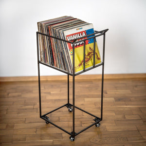 Minimalist black metal vinyl record stand on wheels, filled with colorful LPs, standing on a wooden floor.
