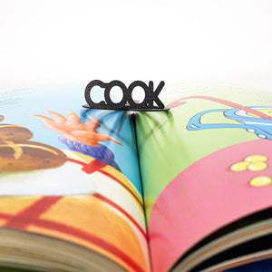 A black metal bookmark with the word “COOK” rests in the center of an open, colorful children’s book with food-themed illustrations.