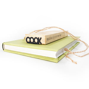 A black metal “COOK” bookmark rests on top of a green hardcover book, alongside a wooden block engraved with the phrase “READING IS GOOD FOR YOU” and tied with twine.