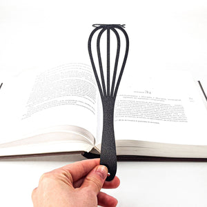 A hand holds a black metal bookmark shaped like a whisk, resting inside an open book.