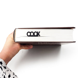 A hand holds a thick closed book with a black metal “COOK” bookmark visible between the pages near the spine.