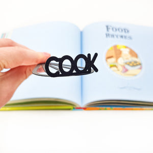 A hand holds a black metal whisk-shaped bookmark with the word “COOK” at the top, in front of an open children’s book titled “Food Rhymes.”