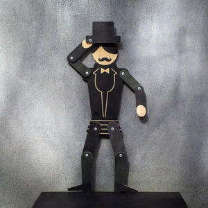 Wooden gentleman puppet on dark shelf against grey wall, tipping top hat with all joints visible