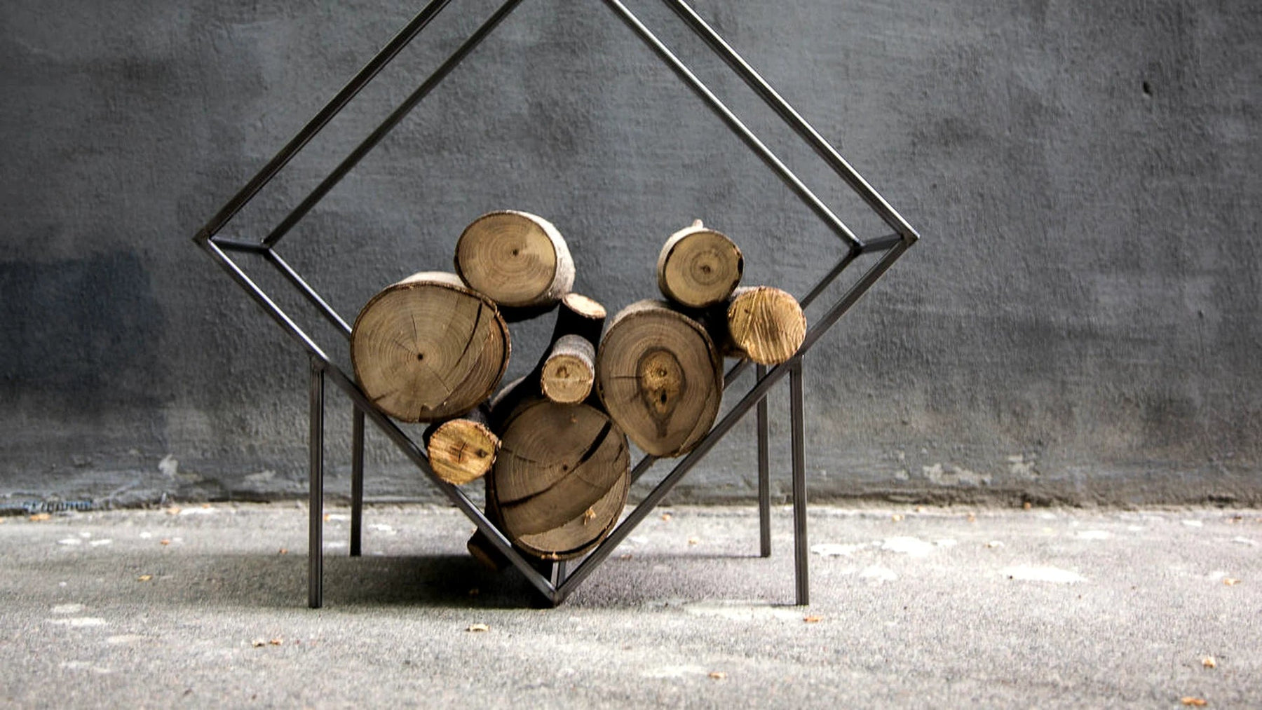 Diamond-shaped steel log holder stacked with split logs against a concrete wall.