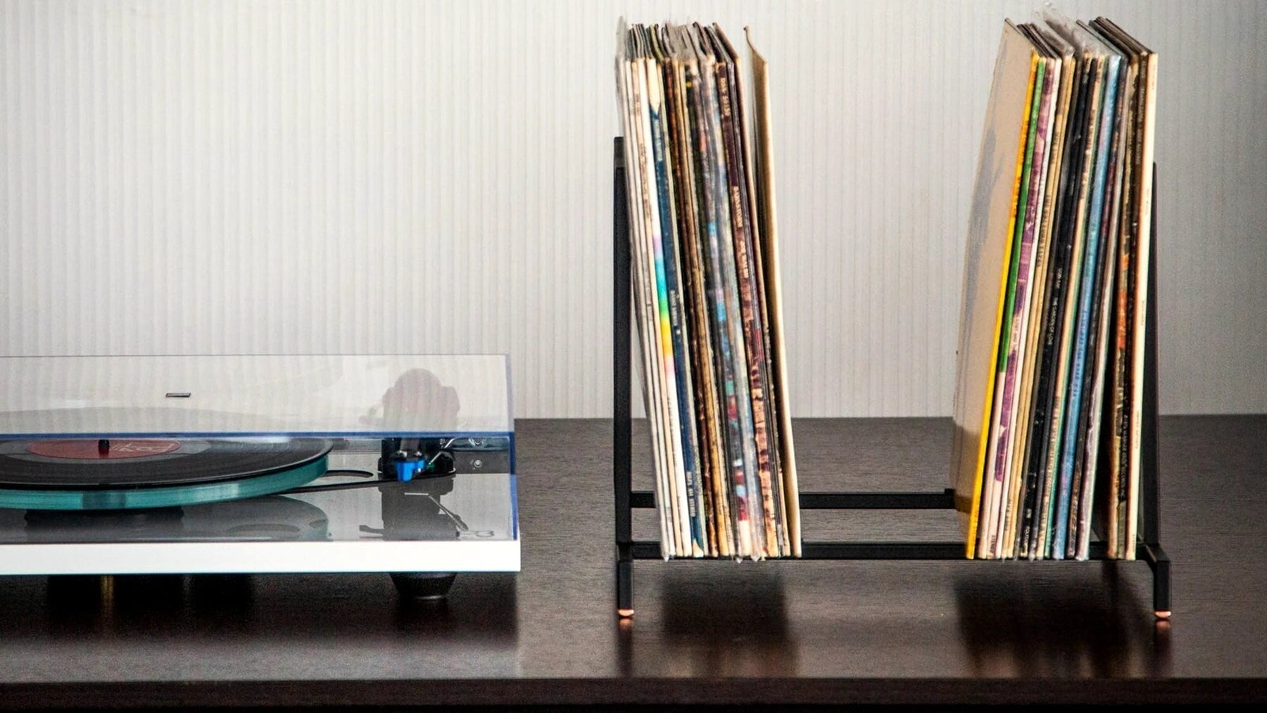 LP records upright in a slim steel stand beside a turntable on a dark shelf.