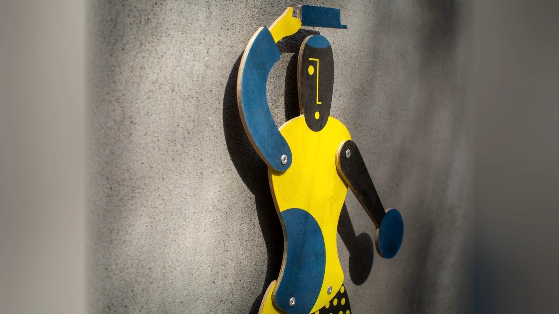 Handmade plywood jumping jack puppet in yellow and blue against a grey wall.