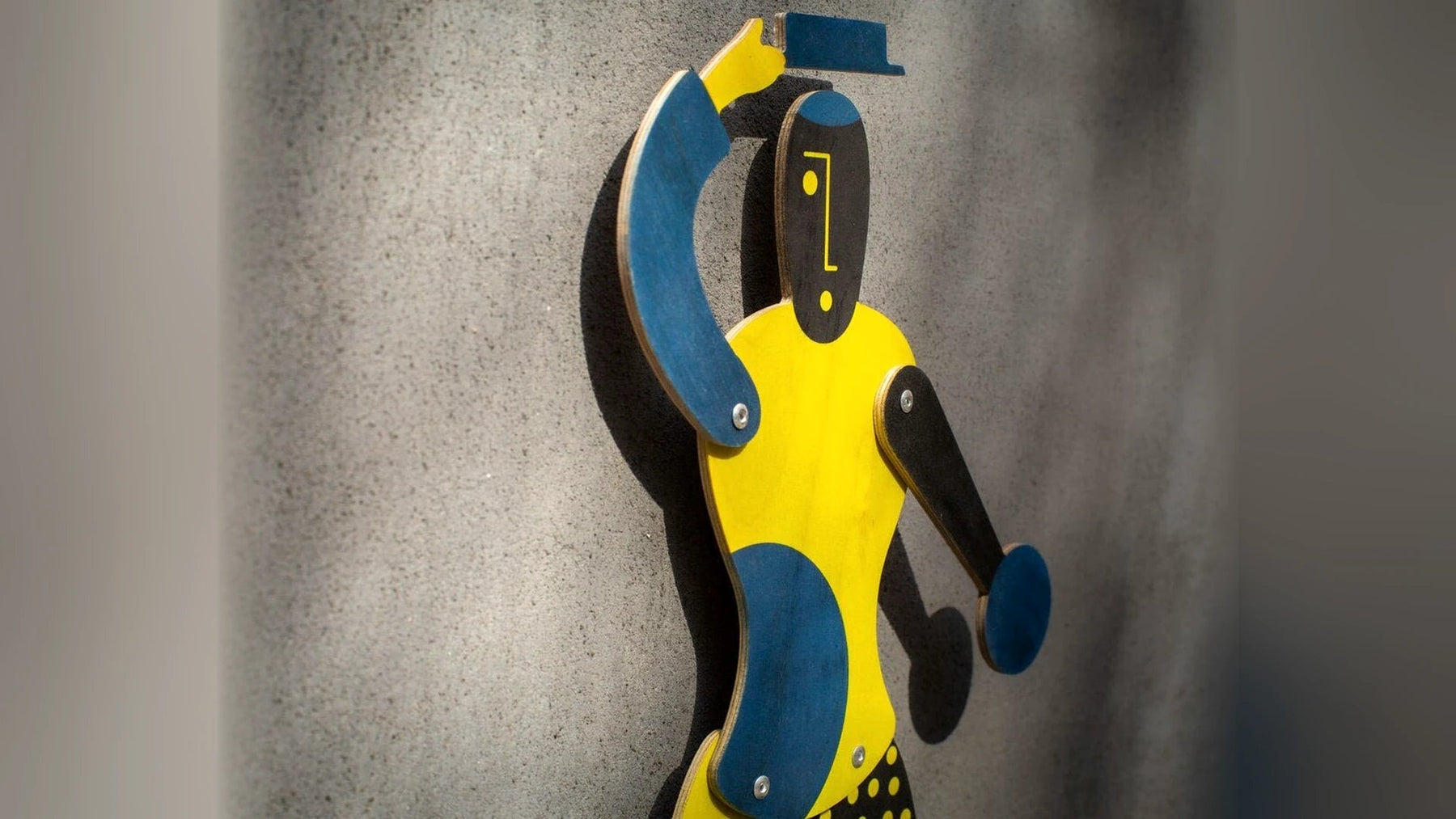 Handmade plywood jumping jack puppet in yellow and blue against a grey wall.