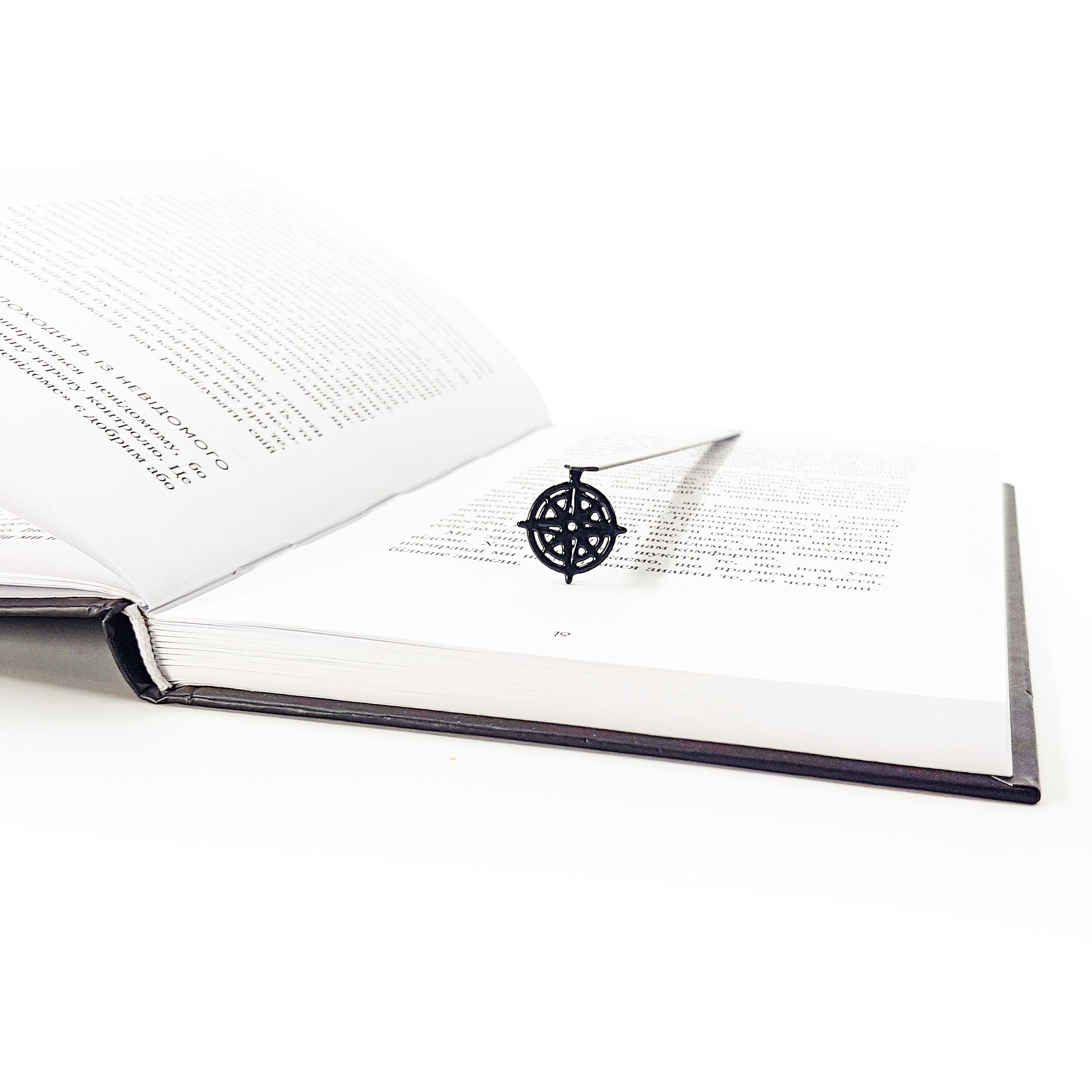 Metal bookmark with compass rose pendant in open dark-bound book, diagonal angle view