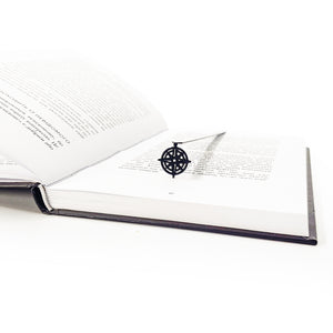 Metal bookmark with compass rose pendant in open dark-bound book, diagonal angle view
