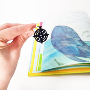 Hand holding compass rose metal bookmark against open colorful children's book