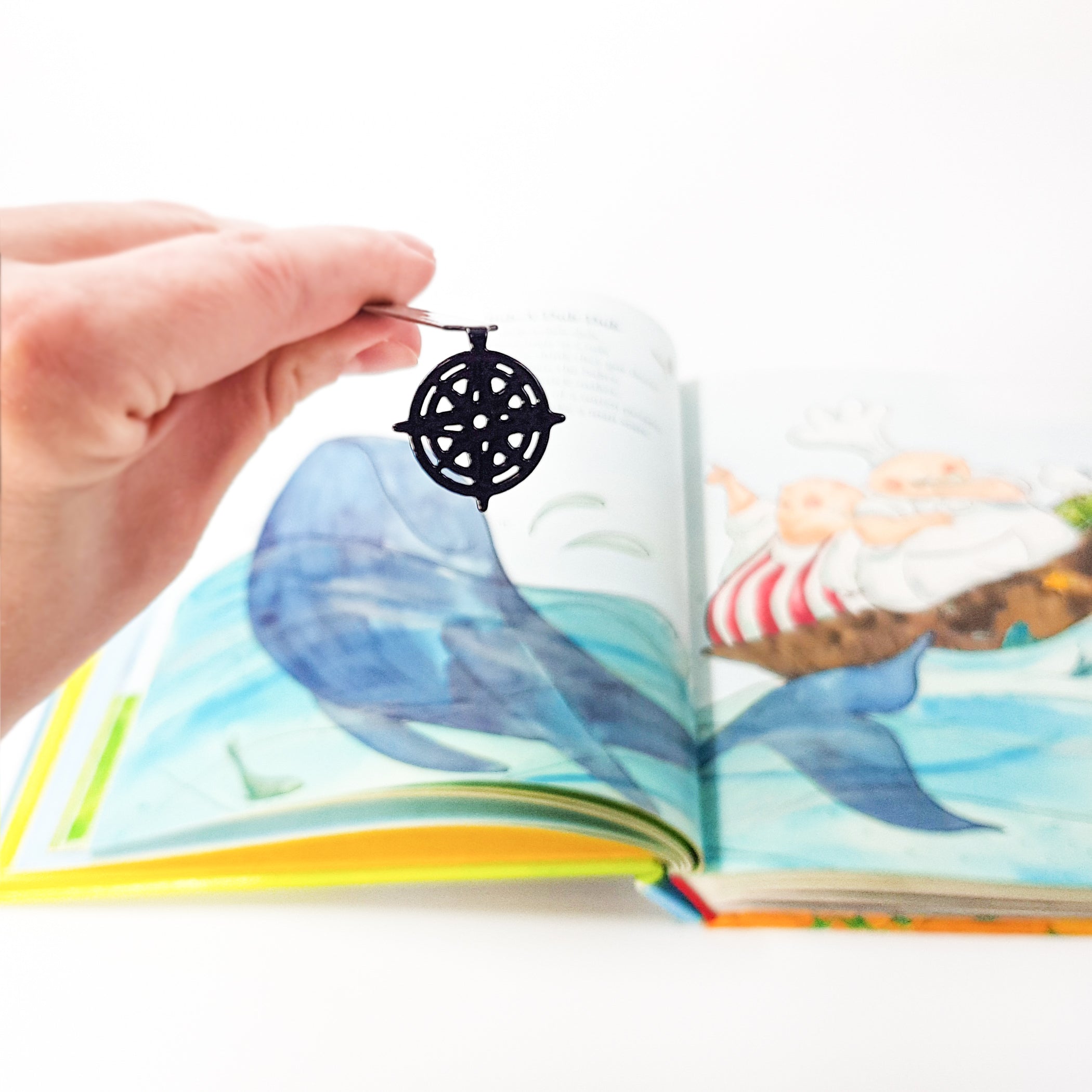 Hand holding black metal compass rose bookmark over open book with whale illustration, full pendant detail