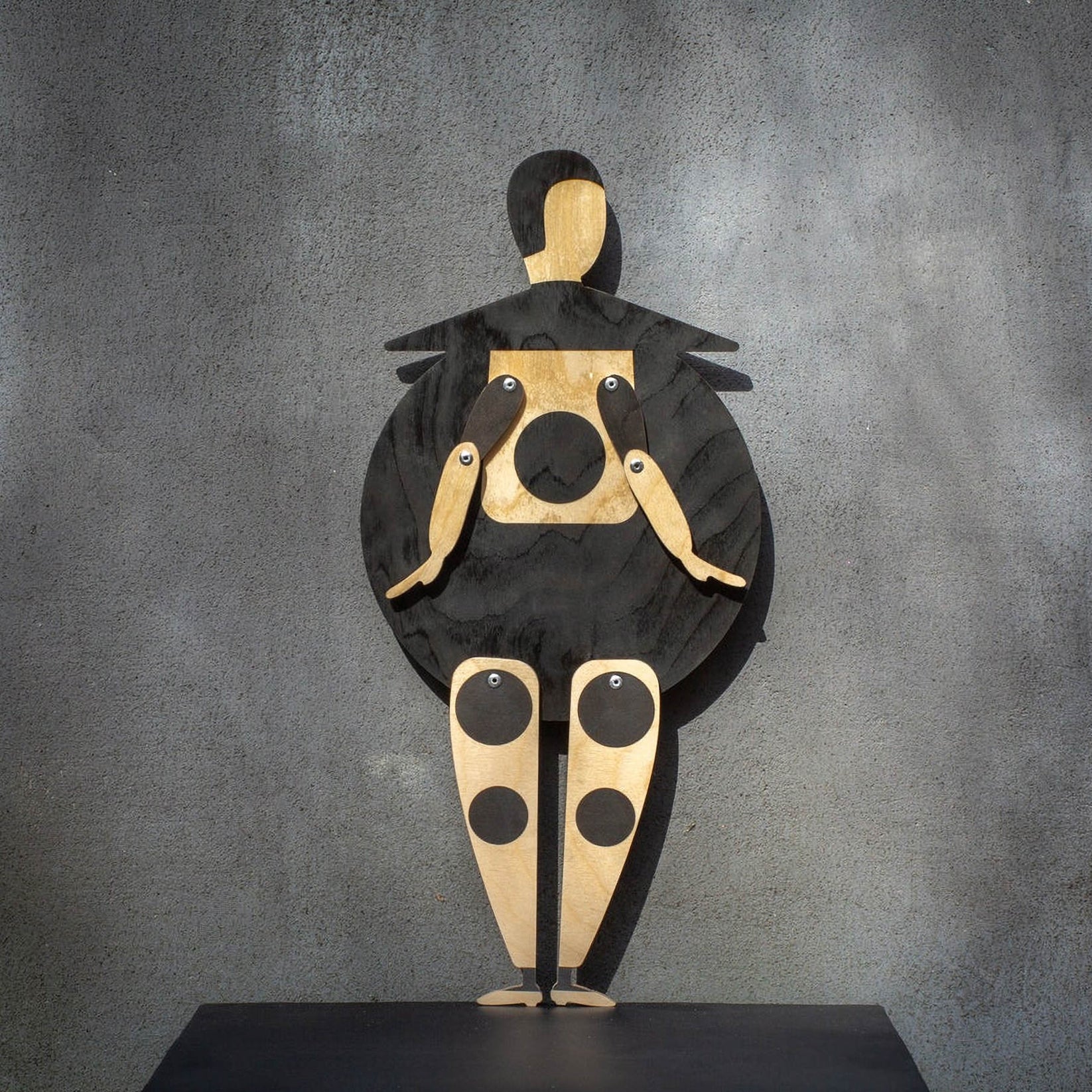 Bauhaus Beetle wooden marionette puppet standing on a black shelf against a grey wall, front view