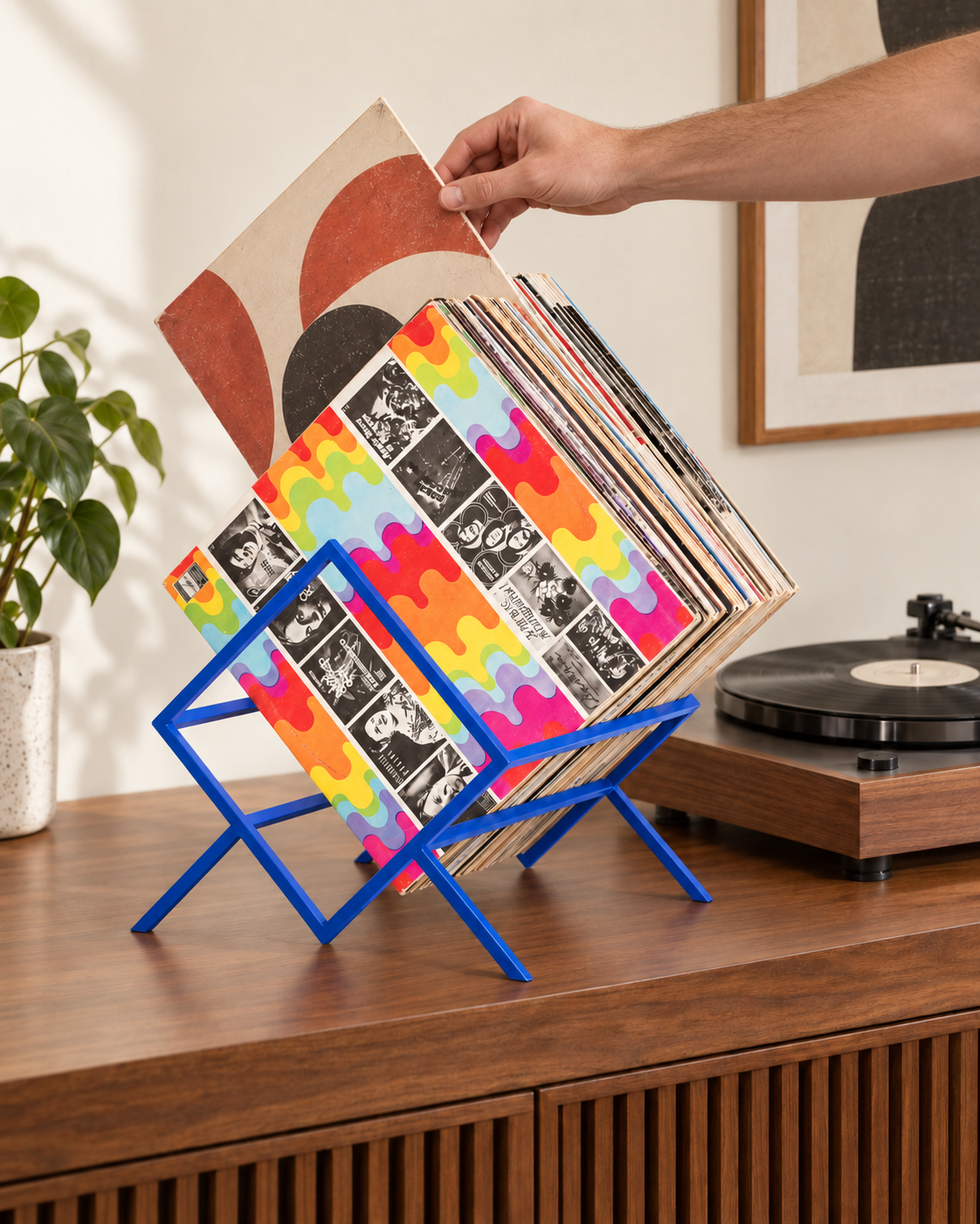 Hand reaching from the turntable side to select a vinyl record from a blue Atelier Article 1Romb stand on a walnut media console.