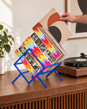 Hand pulling a vinyl record toward the turntable side from a blue Atelier Article 1Romb stand on a walnut media console.