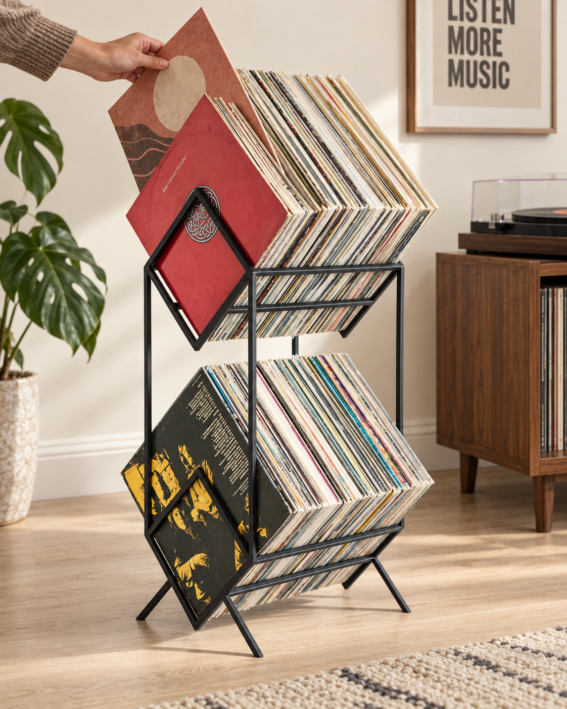 Hand selecting a vinyl record from the upper tier of a black Atelier Article 2Romb stand, with LP sleeves resting diagonally in diamond frames beside a turntable.