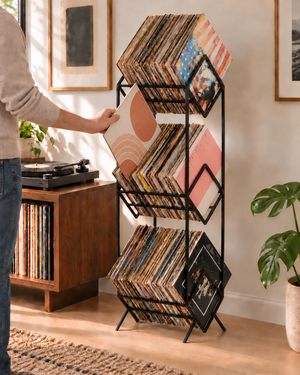 Person taking a vinyl record from the middle tier of a black Atelier Article 3Romb stand beside a turntable.