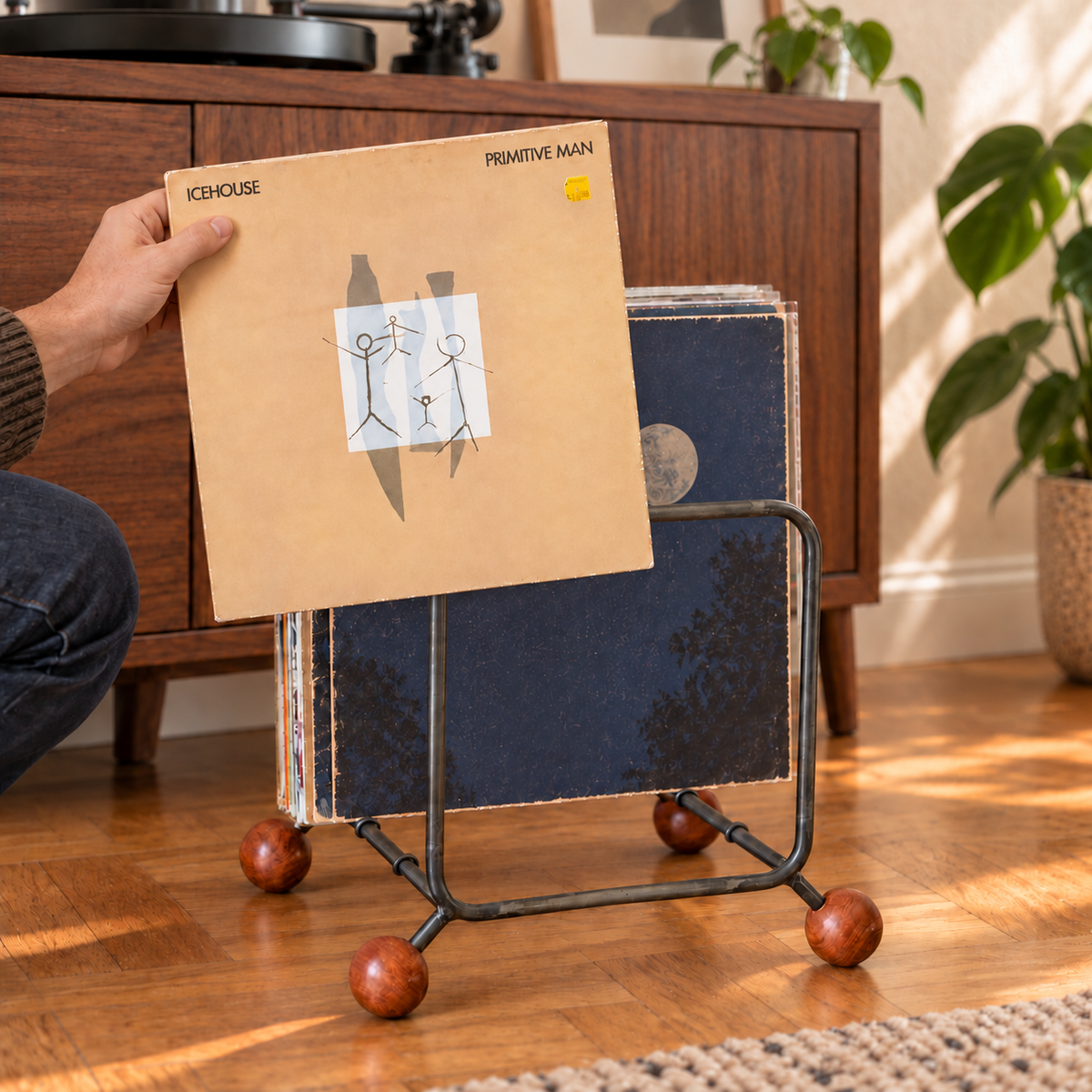 A person holds an Icehouse LP sleeve in front of an Atelier Article steel record rack with four wooden ball feet beside a walnut turntable cabinet.