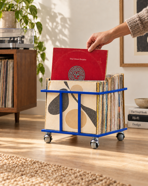 Hand selecting a vinyl record from a blue Atelier Article metal record crate on swivel wheels beside a turntable.