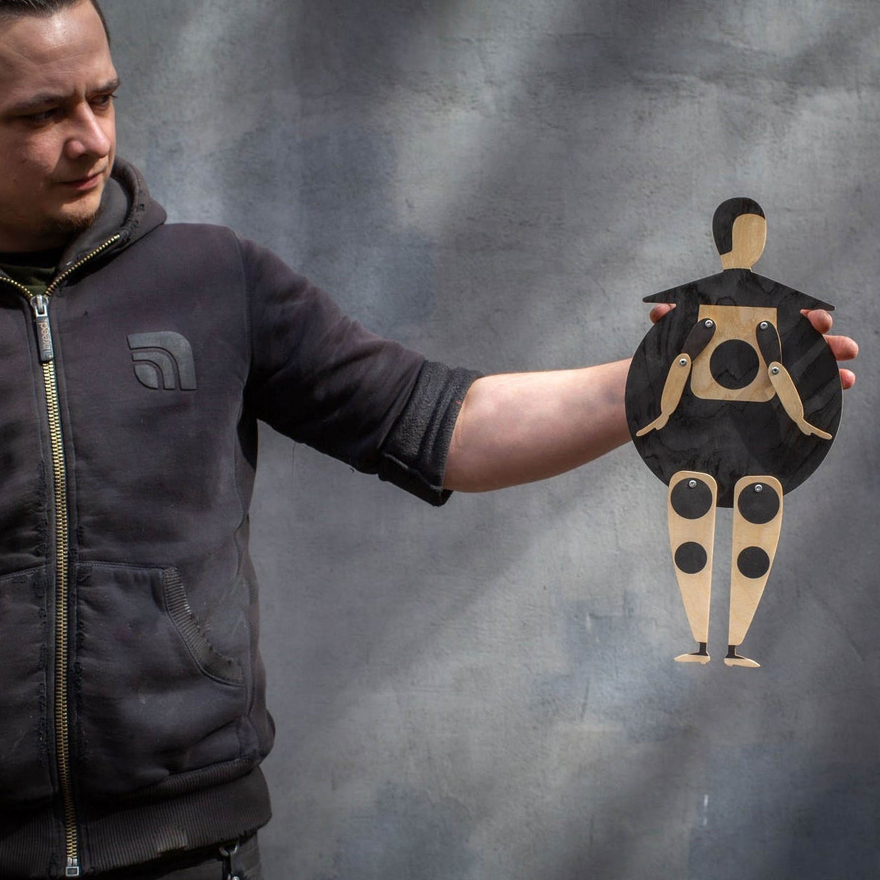 Maker holding the Bauhaus Beetle jumping jack puppet at arm's length against a grey wall, showing its 41 cm size