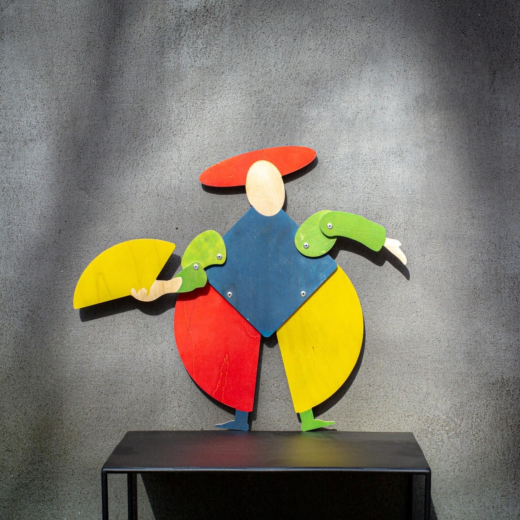 Colorful Bauhaus harlequin wooden puppet displayed on black metal shelf against grey textured wall