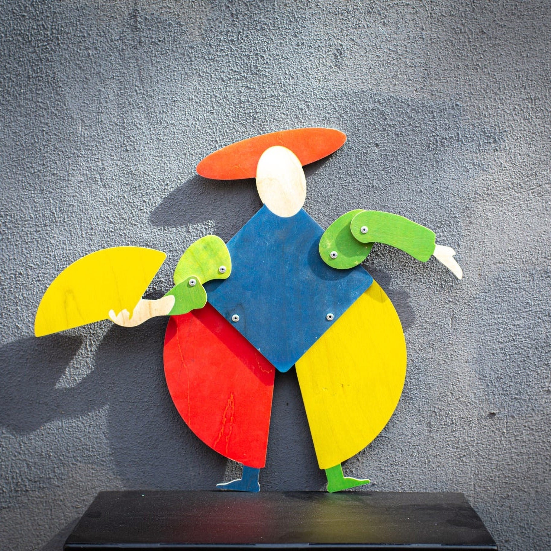 Bauhaus harlequin wooden puppet displayed on textured grey wall, arms posed outward