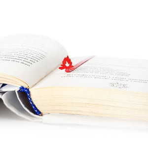 Red bonfire metal bookmark resting inside open book, slim steel page marker with flame shape on printed pages