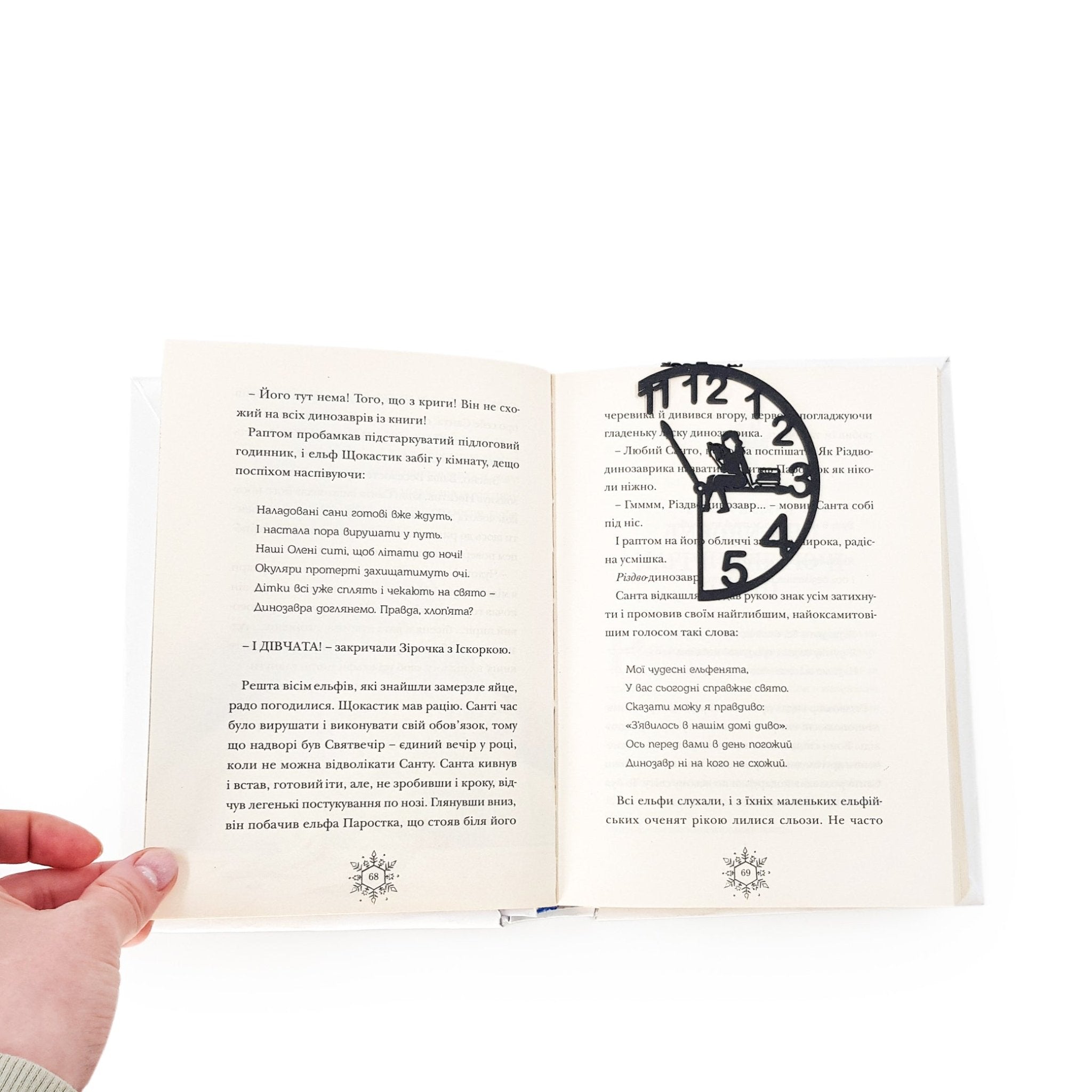 Black metal Book O’Clock bookmark clipped onto an open book, showing the clock-shaped design with a reading figure marking the page on a white background.
