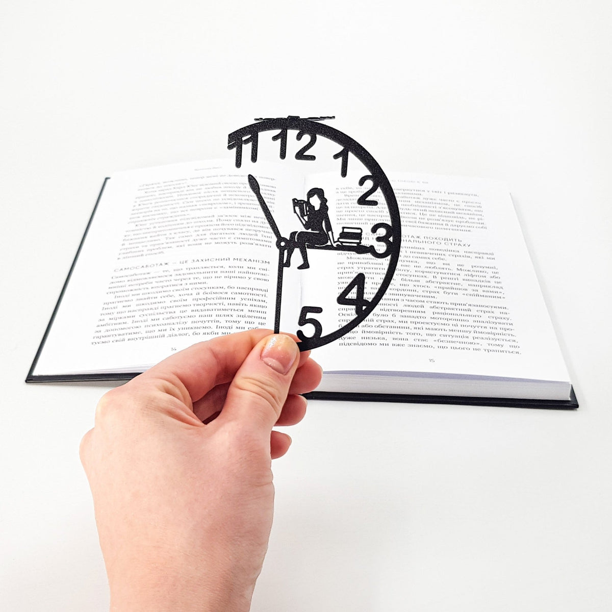 Hand holding a black metal Book O’Clock bookmark with a clock-shaped design and a reading figure, placed over an open book on a white background.