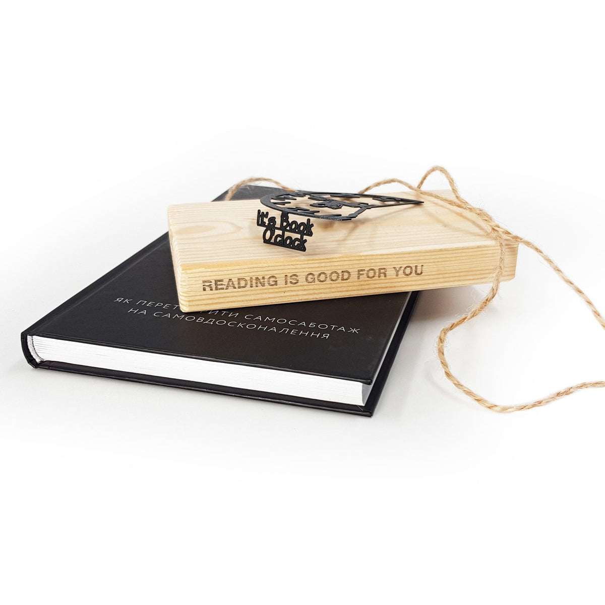 Black metal Book O’Clock bookmark resting on a wooden block engraved “Reading is good for you,” placed on top of a closed black book with natural twine packaging on a white background.
