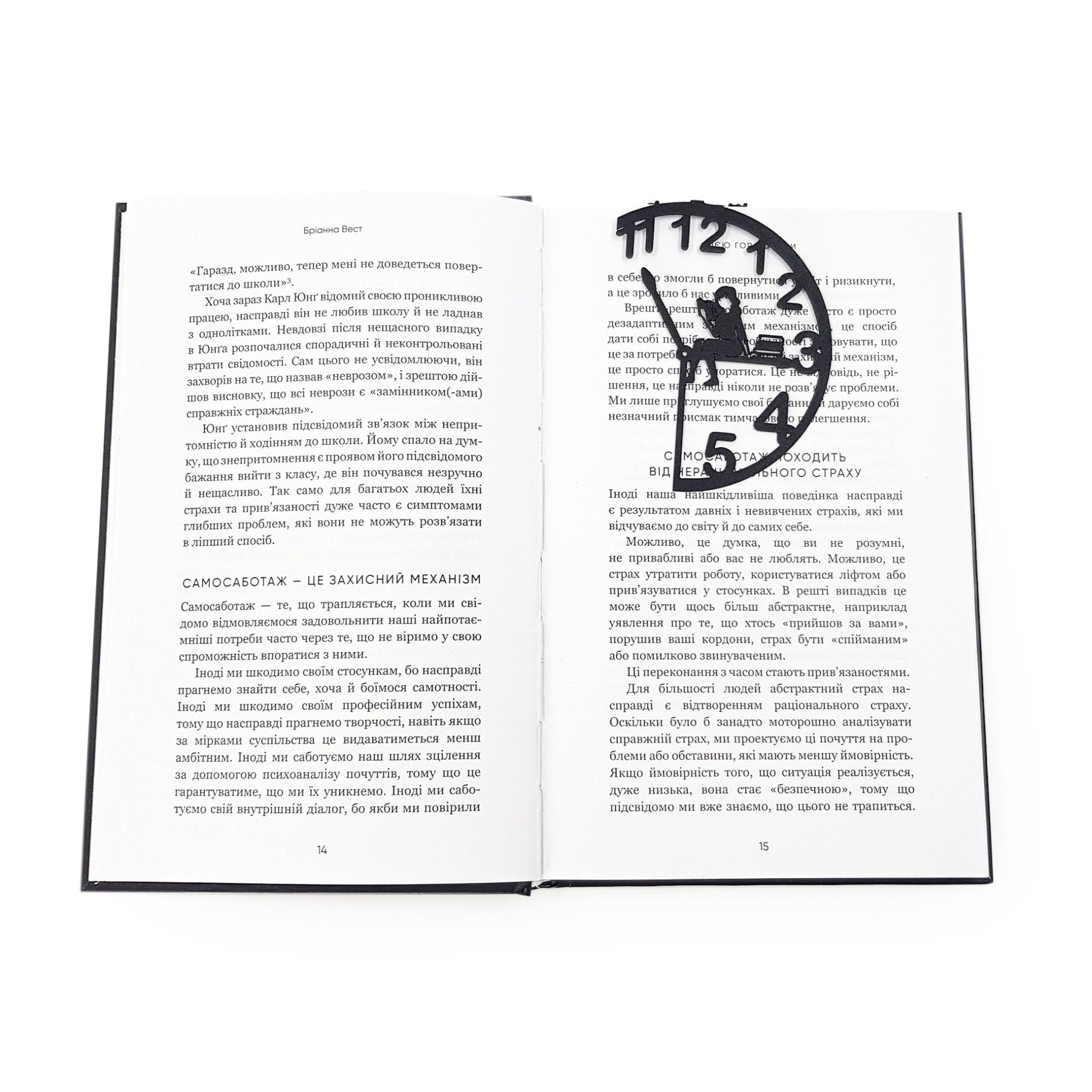 Black metal Book O’Clock bookmark marking a page in an open book, showing the clock-shaped design with a seated reading figure against a white background.