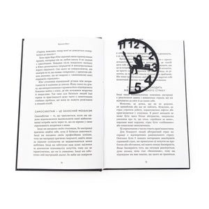 Black metal Book O’Clock bookmark marking a page in an open book, showing the clock-shaped design with a seated reading figure against a white background.
