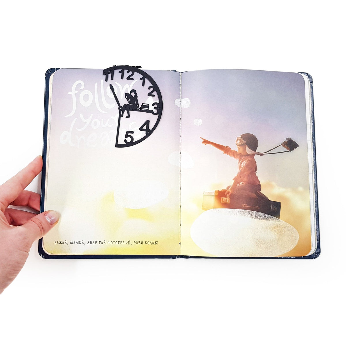 Black metal Book O’Clock bookmark marking a page in an open illustrated book, showing the clock-shaped design with a reading figure against a dreamy, sky-themed spread.
