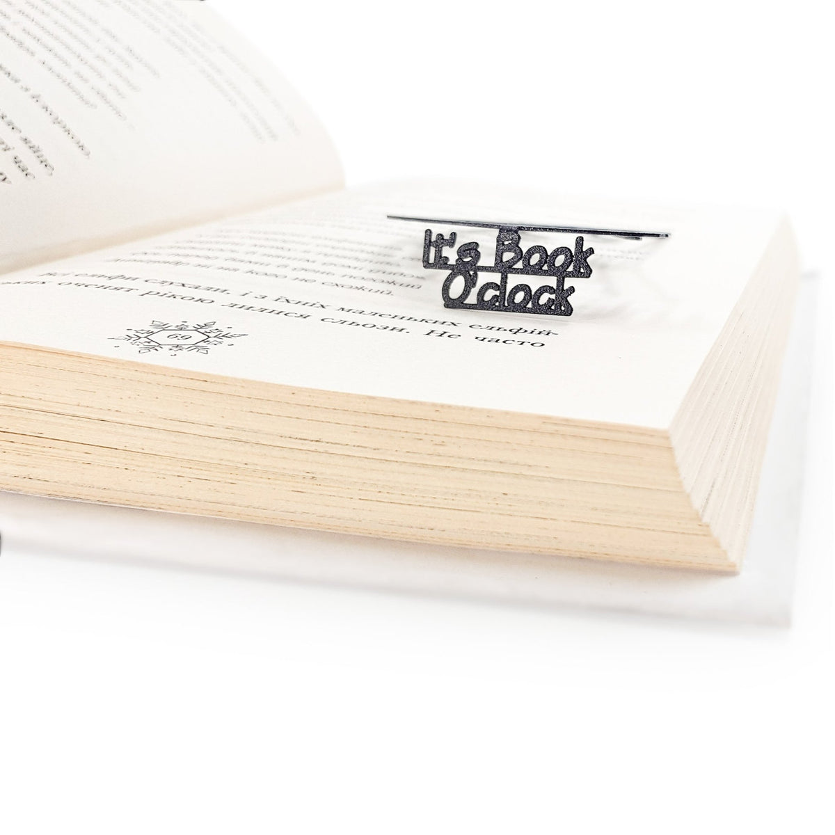 Close-up side view of the black metal Book O’Clock bookmark clipped onto the pages of an open book, showing its thin profile and cut-out text detail on a white background.