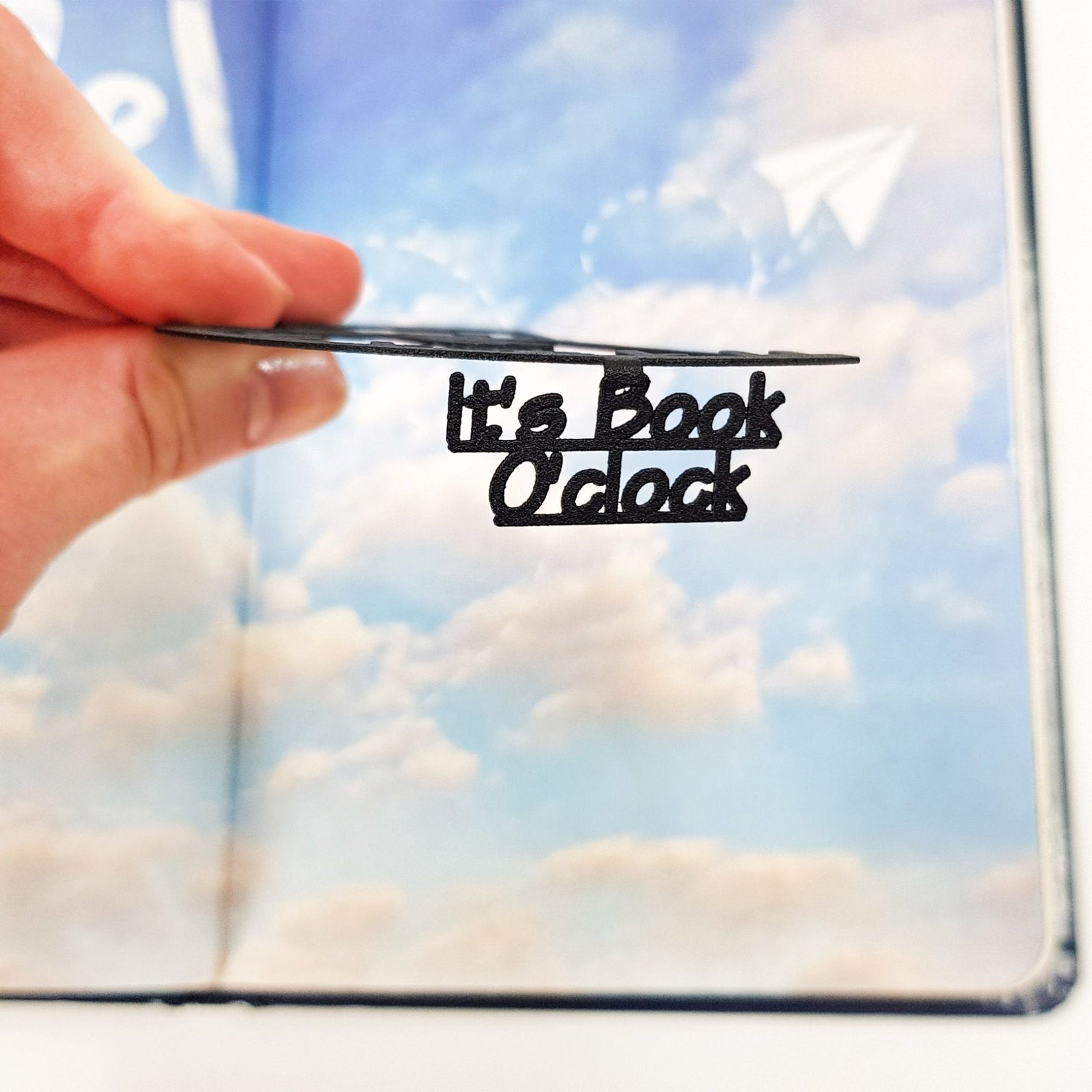 Side view of the black metal Book O’Clock bookmark showing its slim profile and the cut-out “It’s Book O’Clock” text, held over an open book with a sky-patterned page.