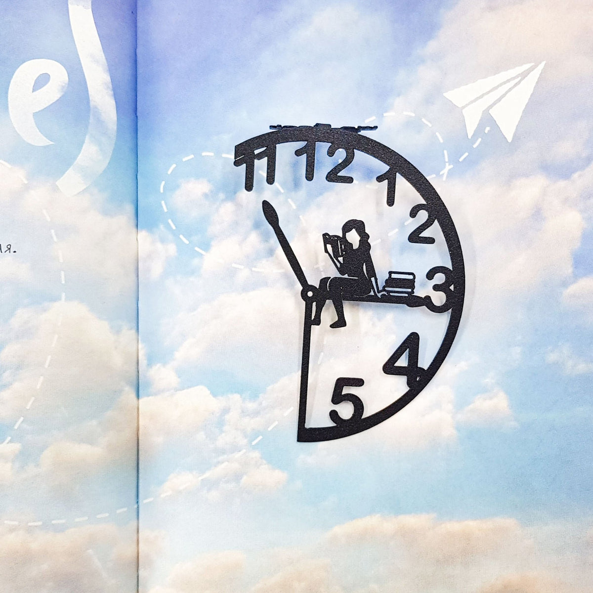 Black metal Book O’Clock bookmark with a clock-shaped design and a seated reading figure, placed on an open book with a sky and clouds illustration in the background.