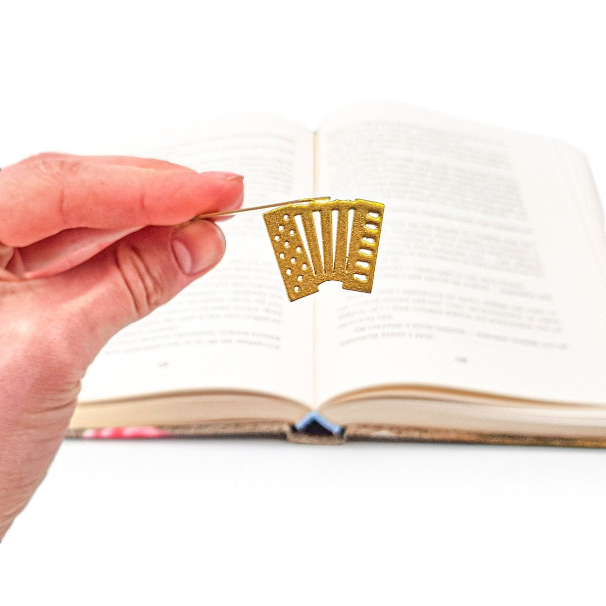Golden Accordion metal bookmark in gold held in a hand above an open book