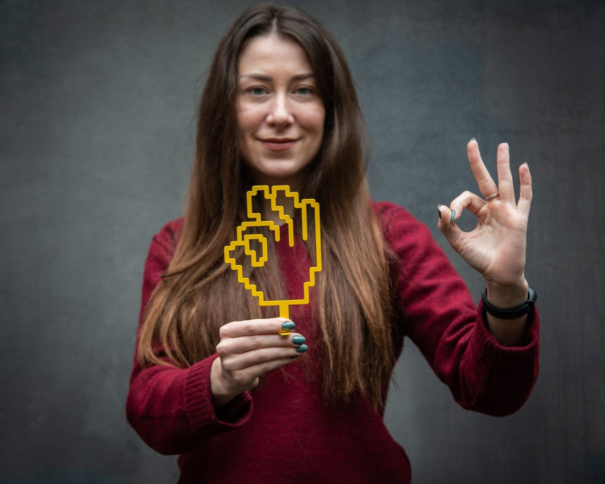 OK Sign Pixel Style metal bookmark in gold held in a hand above an open book