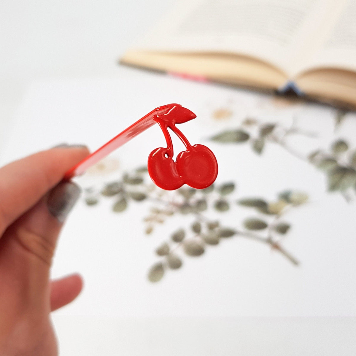 Sweet Cherries metal bookmark in red held up beside an open book