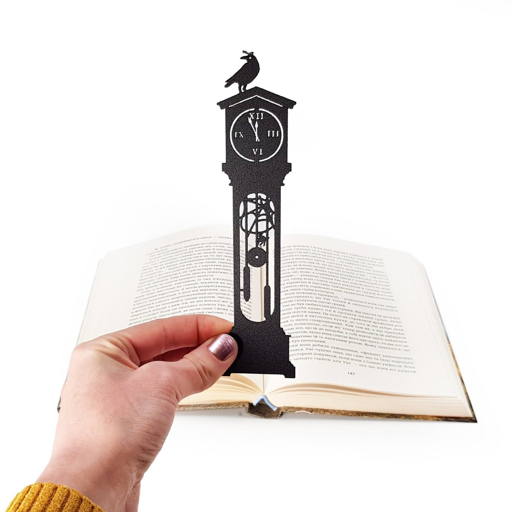 The Raven's Pendulum Dance metal bookmark held in a hand above an open book