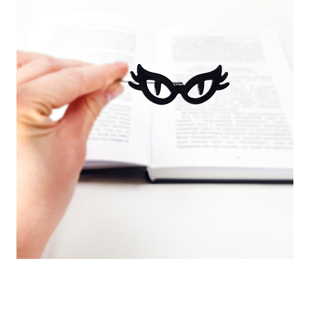 Cat’s Eye Glasses metal bookmark held in a hand above an open book