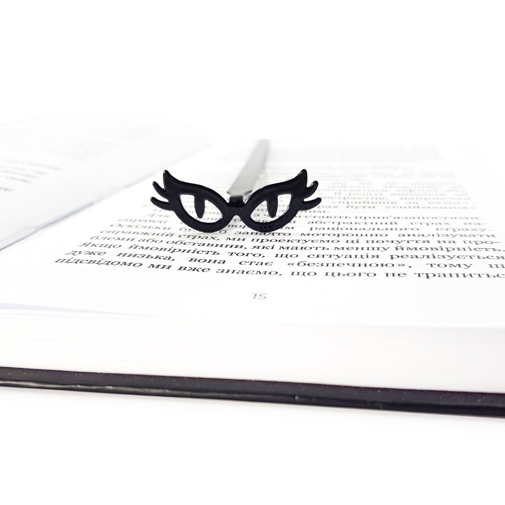Black metal bookmark shaped like cat’s eye glasses resting on an open book, minimalist masquerade-inspired reading accessory for book lovers.
