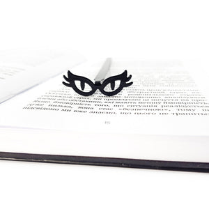 Black metal bookmark shaped like cat’s eye glasses resting on an open book, minimalist masquerade-inspired reading accessory for book lovers.